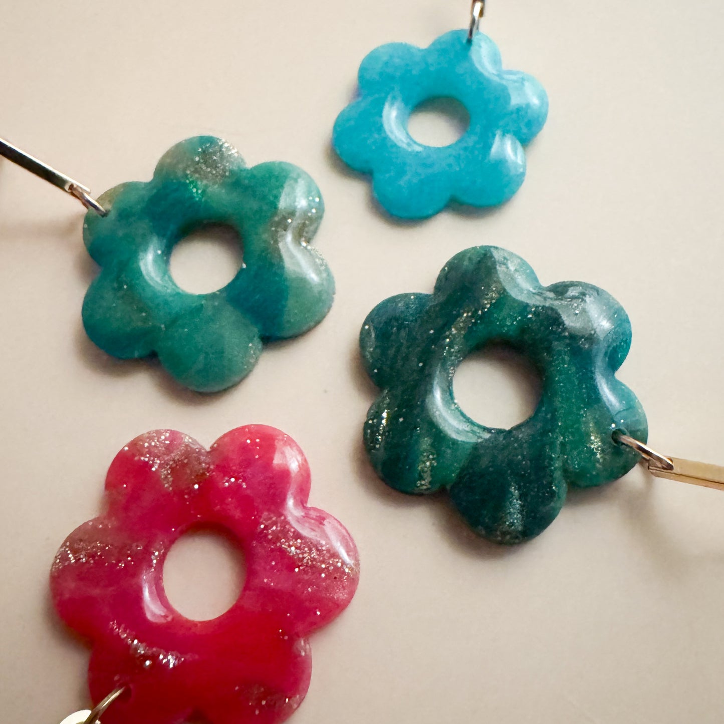 Collection of daisies, one is vibrant pink, a dark teal, and a brighter teal. These all have specks of golden glitter. There is a smaller ice blue daisy as well.