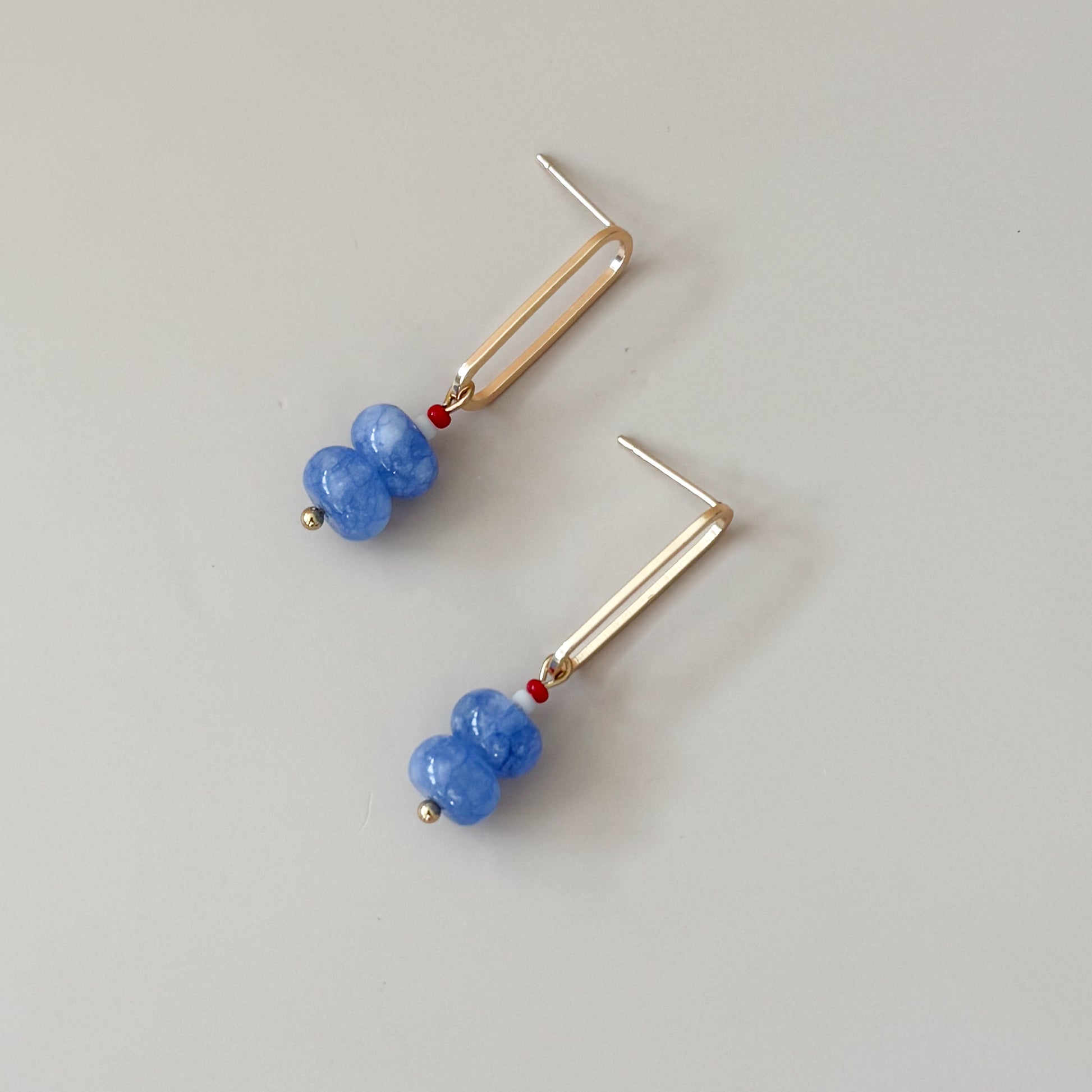 Pair of earrings with blue beads  and smaller red and white beads on top and gold oval on a light gray background